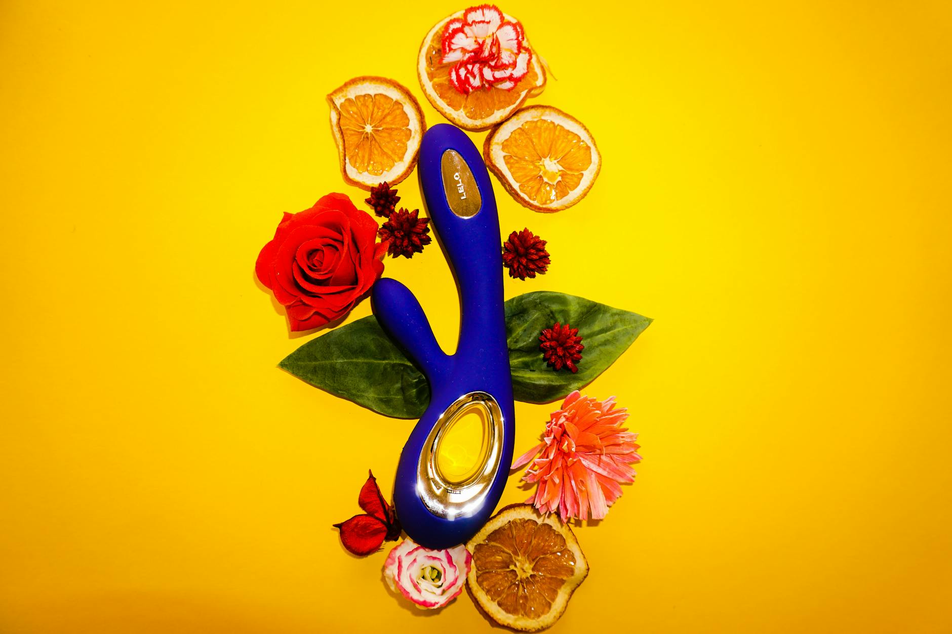 A blue vibrator surrounded by flowers and citrus fruit on a yellow background.